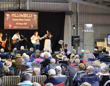 People sitting watching a performance on a stage with a sign saying 'Yellow belly Festival' behind them